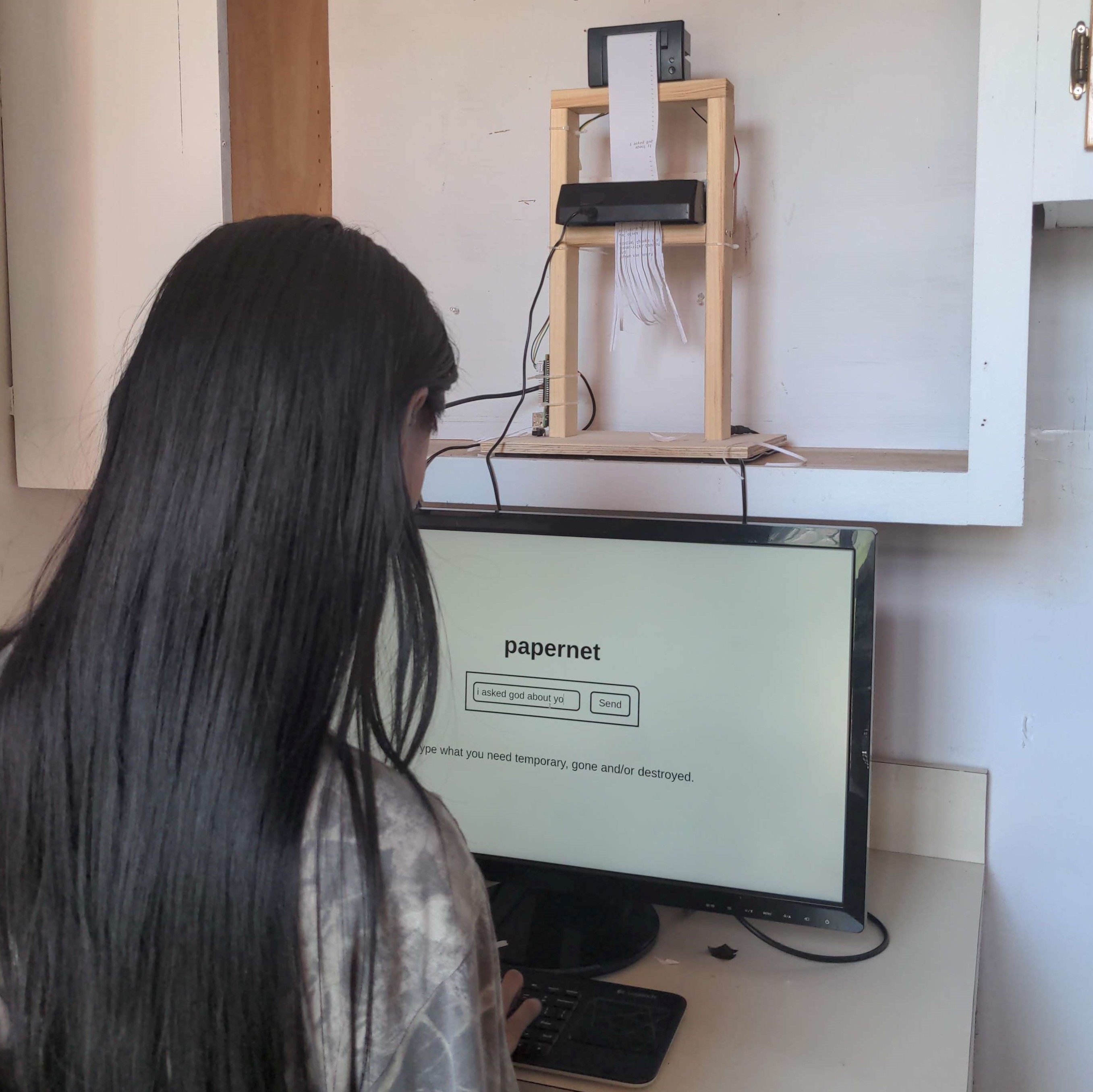 Photo of a robotic mechanism in wooden casing with a small printer atop and a small paper shredder below, paper coming out of the printer is fed into the shredder and shown shredded below.